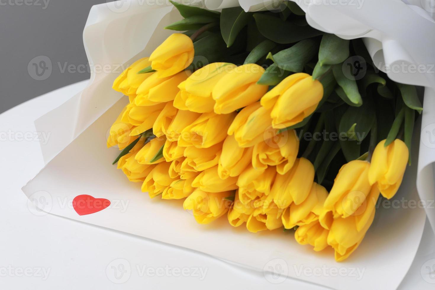 beau gros bouquet de tulipes jaunes dans un emballage blanc et des confettis coeur rouge sur table blanche se bouchent. félicitation. fond festif. espace de copie. photo