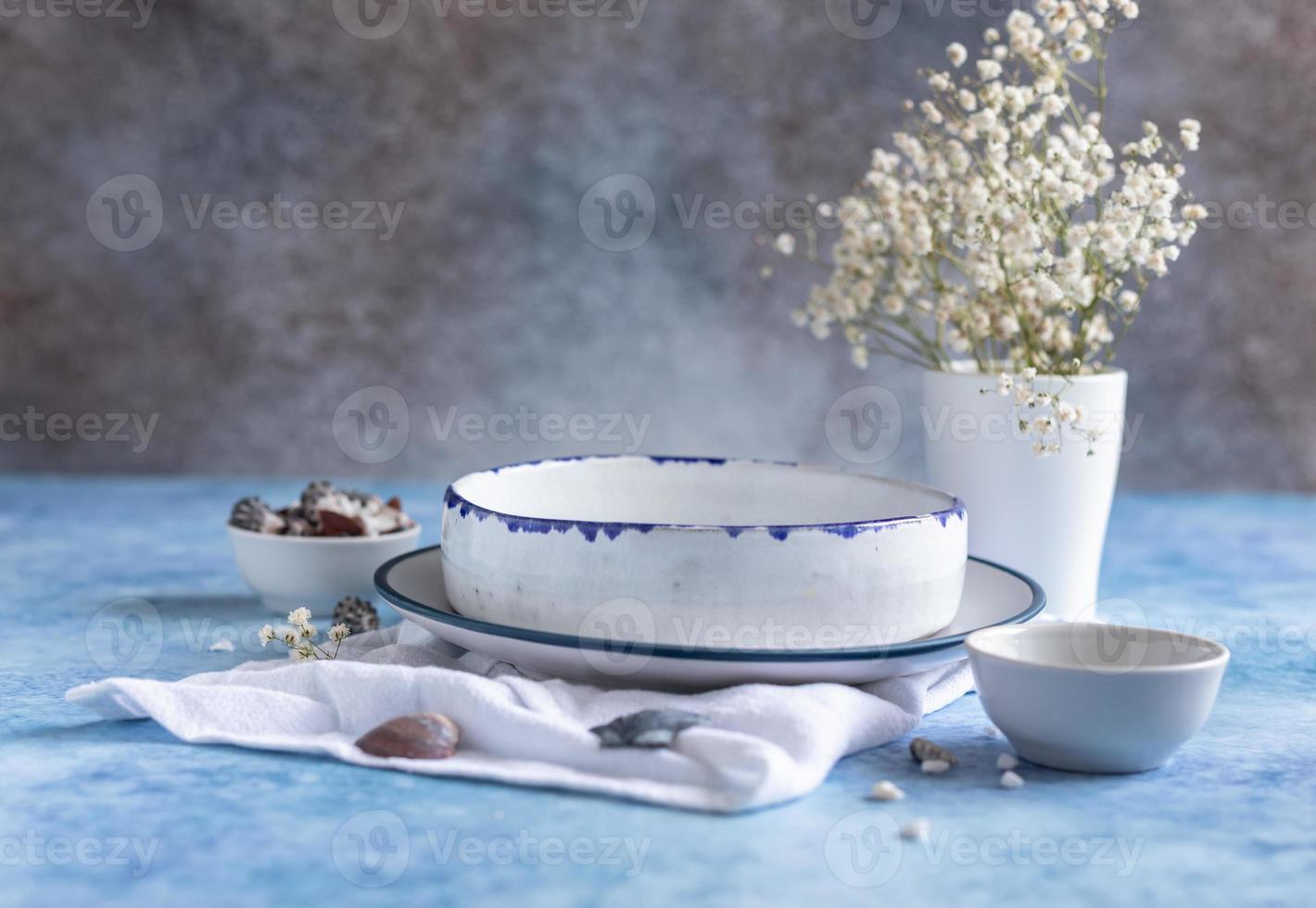 assiettes blanches en céramique faites à la main avec une bande bleue sur le bord, des fleurs séchées et des coquillages vides sur fond de béton bleu. photo