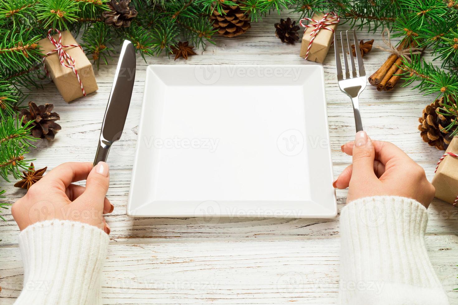 vue en perspective fille tient une fourchette et un couteau à la main et est prête à manger. assiette carrée blanche vide sur fond de noël en bois. concept de plat de dîner de vacances avec un décor de nouvel an photo