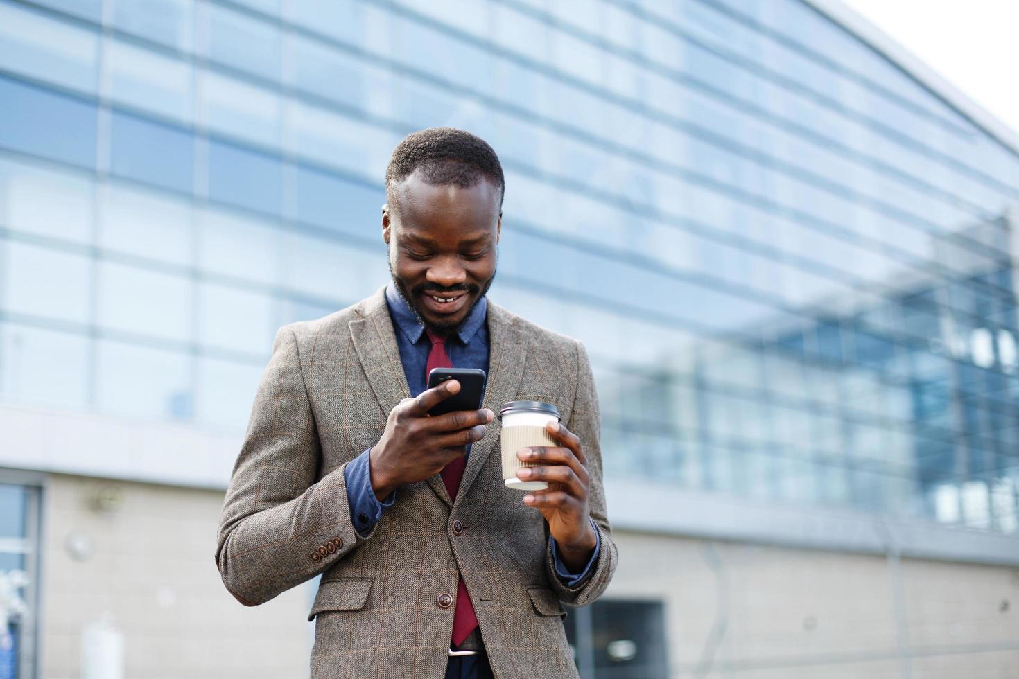 Heureux homme afro-américain lisant quelque chose dans son smartphone alors qu'il se tient à l'extérieur avec une tasse de café photo