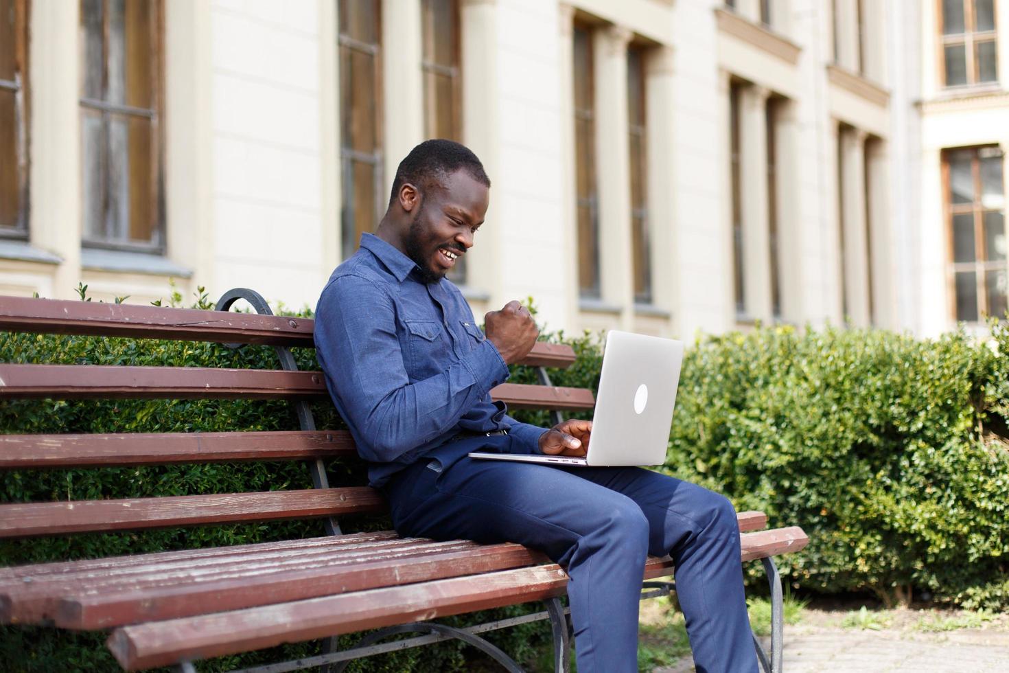 Heureux homme afro-américain travaille sur son ordinateur portable assis sur le banc à l'extérieur photo
