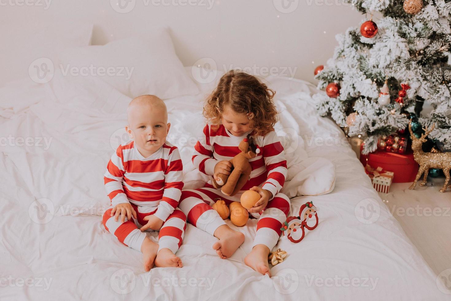 les enfants en pyjama rouge et blanc essaient des lunettes amusantes avec le père noël assis dans son lit. mode de vie. frère et soeur célébrant noël. garçon et fille jouent à la maison. photo de haute qualité