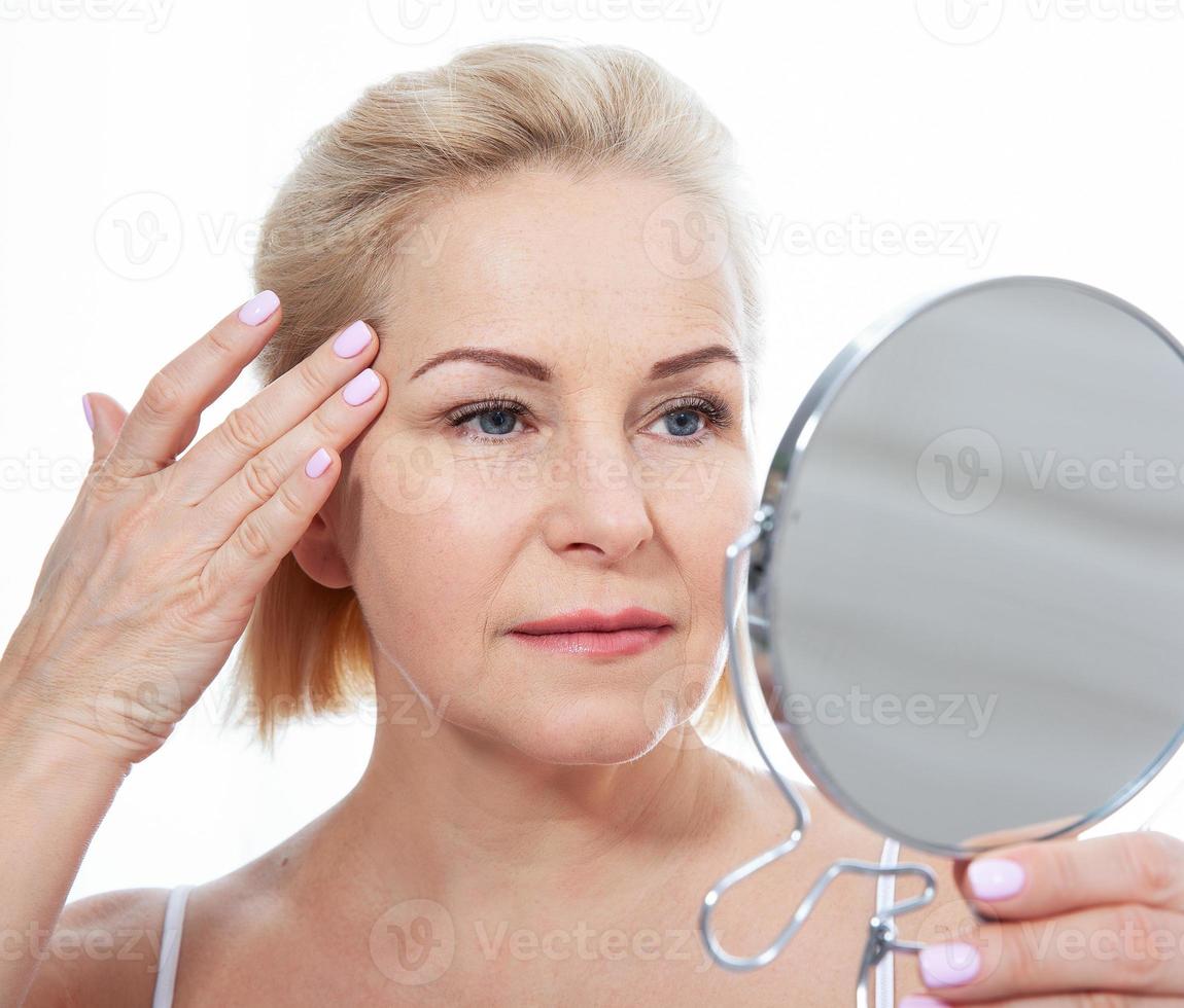 heureux modèle de femme d'âge moyen touchant la peau du visage regardant dans le miroir. dame âgée mature souriante bénéficiant de soins de la peau sains, beauté vieillissante, concept de produits cosmétiques de traitement de soins de la peau. photo