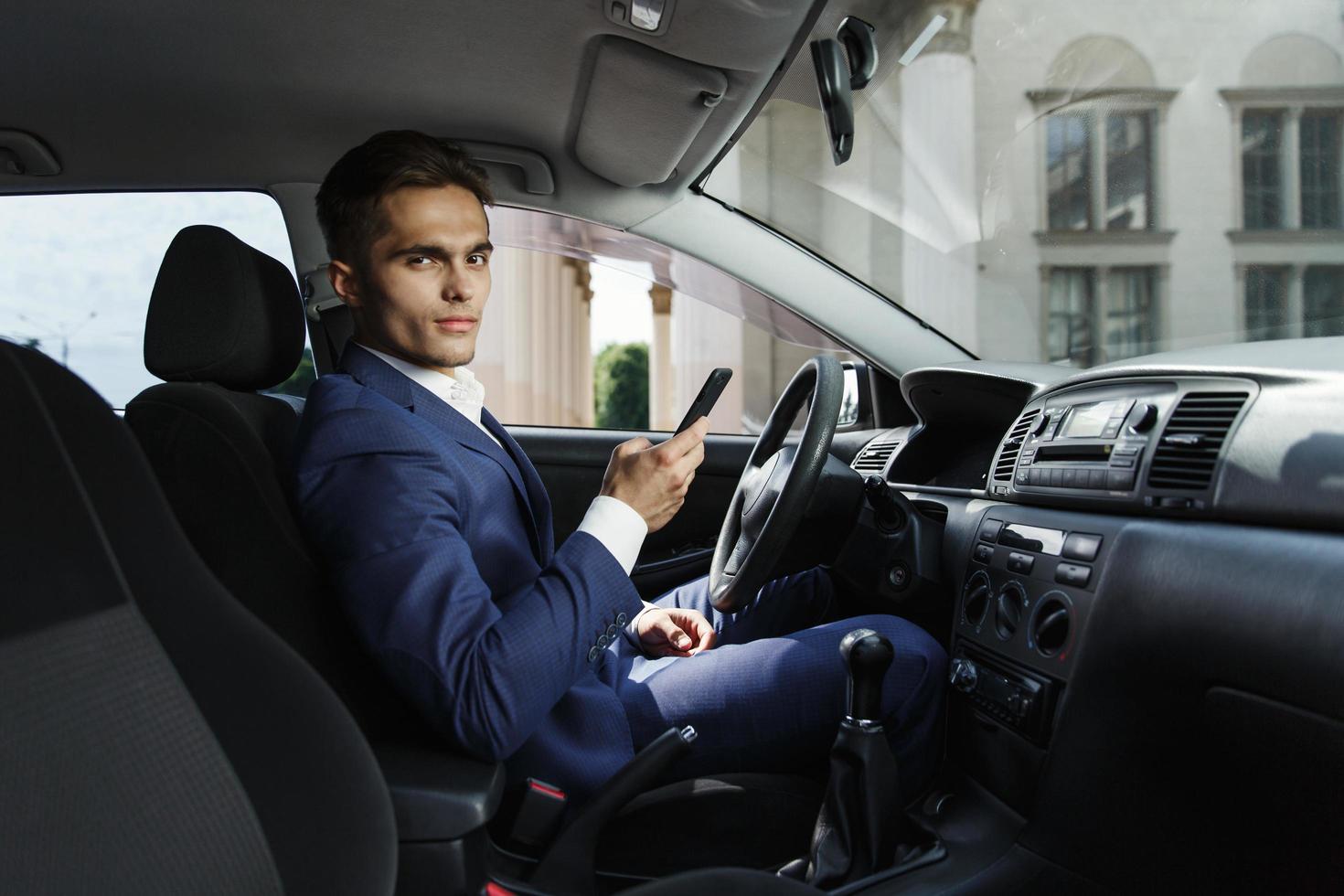 homme d'affaires souriant est assis à l'intérieur de la voiture et travaille avec son smartphone photo