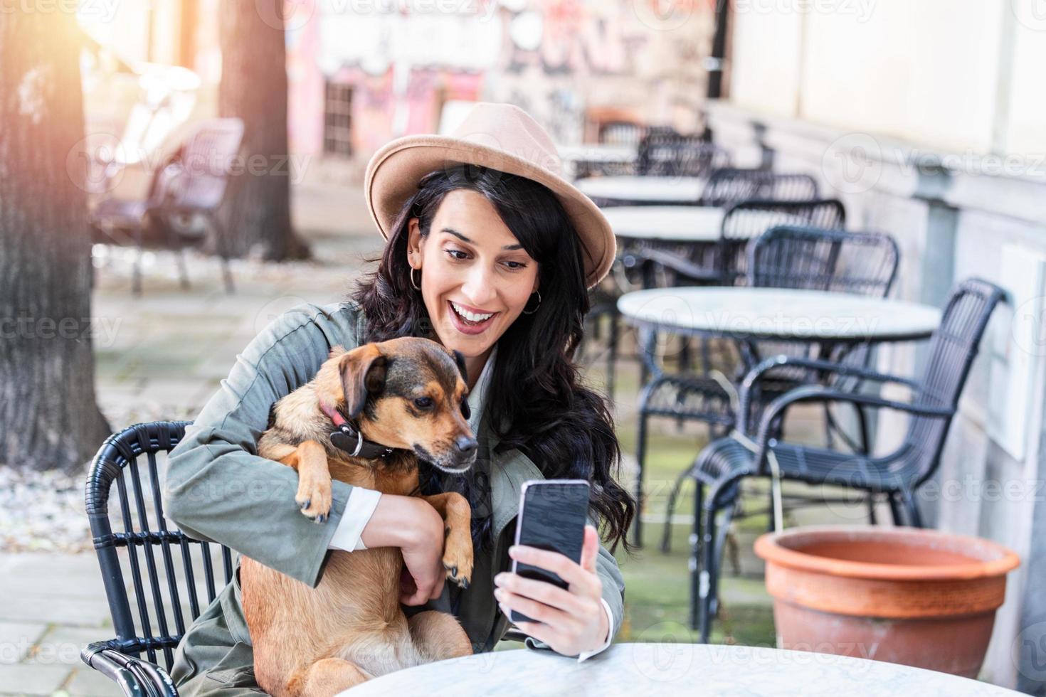 jeune femme avec son chien dans une cafétéria acceptant les animaux. fille buvant du café au café local avec son chien prenant un selfie avec un téléphone portable photo