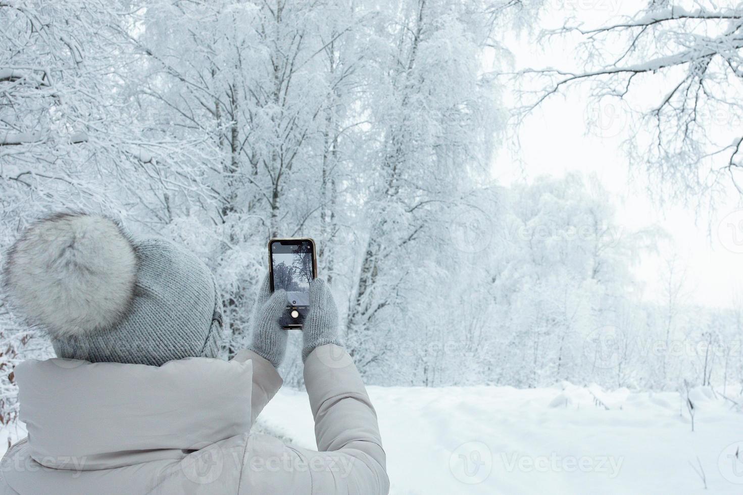 femme en bonnet gris tricoté avec pompon de derrière prenant une photo de paysage de forêt d'hiver neigeant avec son smartphone