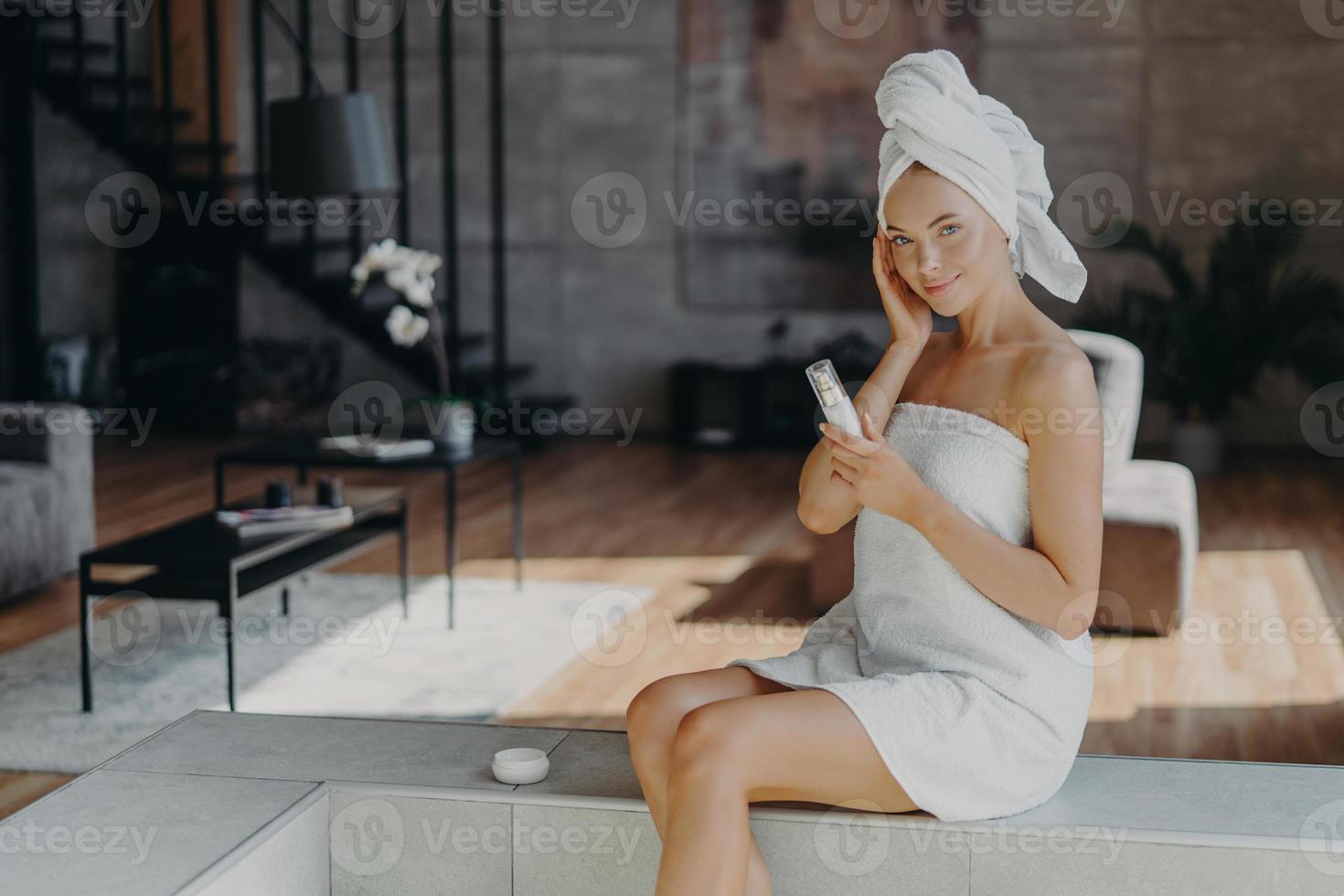 une femme en bonne santé et heureuse applique une lotion pour le corps et une crème pour le visage, nourrit la peau, enveloppée dans une serviette de bain, sourit doucement, pose contre l'intérieur domestique, subit des soins de beauté. notion de dermatologie photo