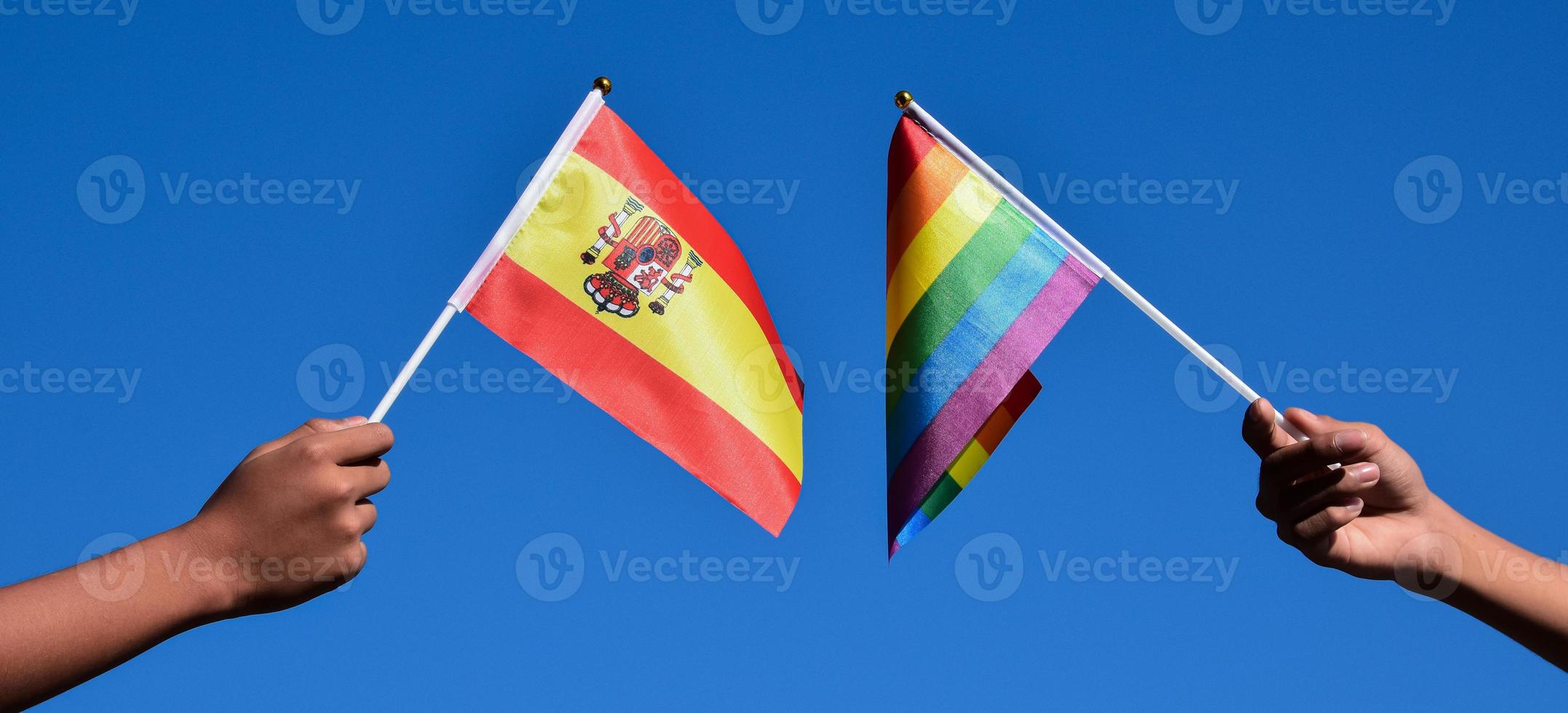 drapeau espagnol et drapeau arc-en-ciel, symbole lgbt, tenant dans les mains, fond bleu ciel, concept pour la célébration lgbt en espagne et dans le monde pendant le mois de la fierté, juin, mise au point douce et sélective, espace de copie. photo