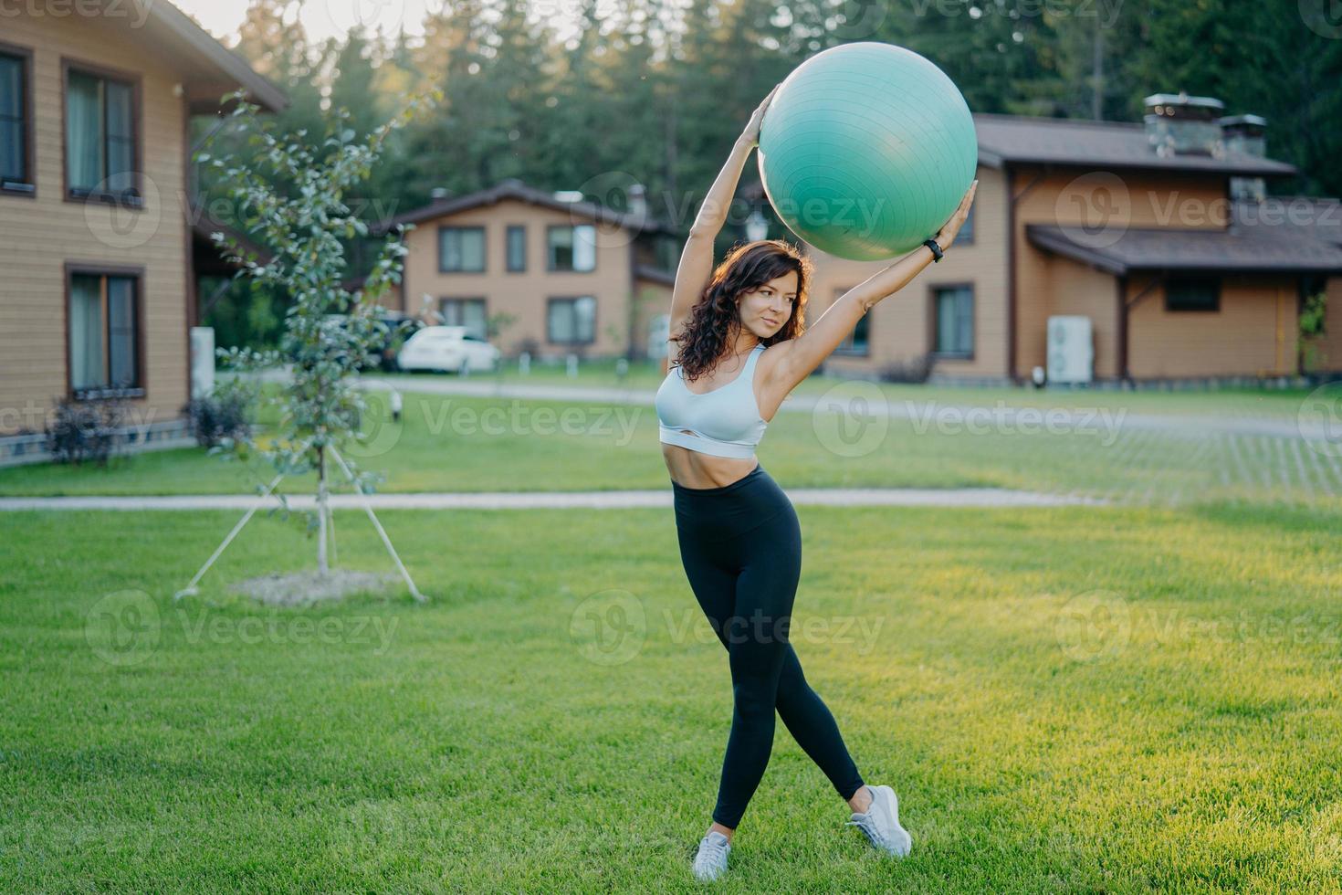 photo pleine longueur d'une femme brune mince et sérieuse a une forme de corps parfaite tient un ballon de fitness au-dessus de la tête, fait des exercices de fitness en plein air près de la maison, vêtue d'un haut court, de leggings et de baskets.