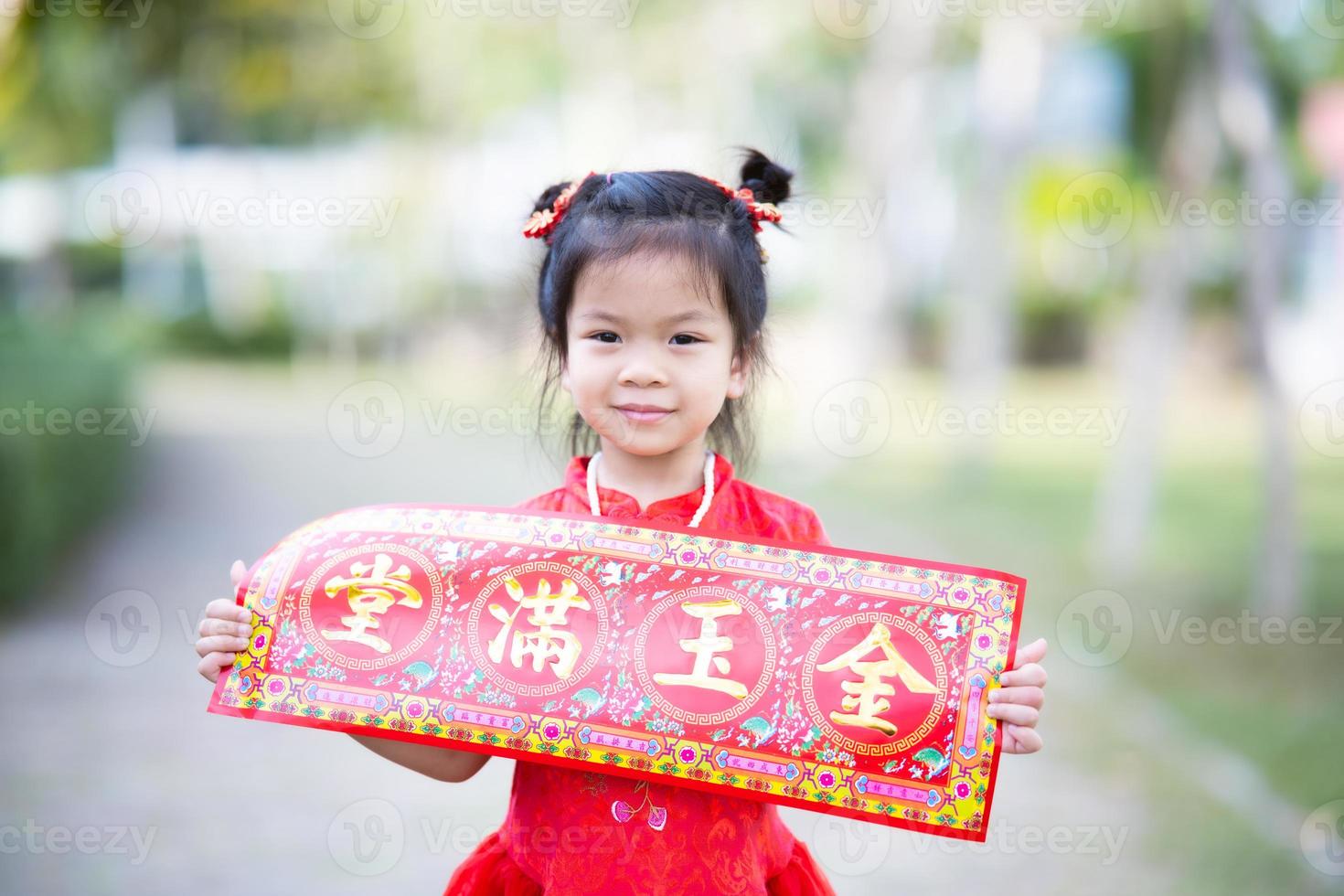 portrait jolie fille asiatique tenant une carte de voeux de nouvel an chinois qui signifie riche et heureux. les enfants sourient doucement. l'enfant porte du cheongsam rouge dans le jardin. enfant de 5 ans. photo