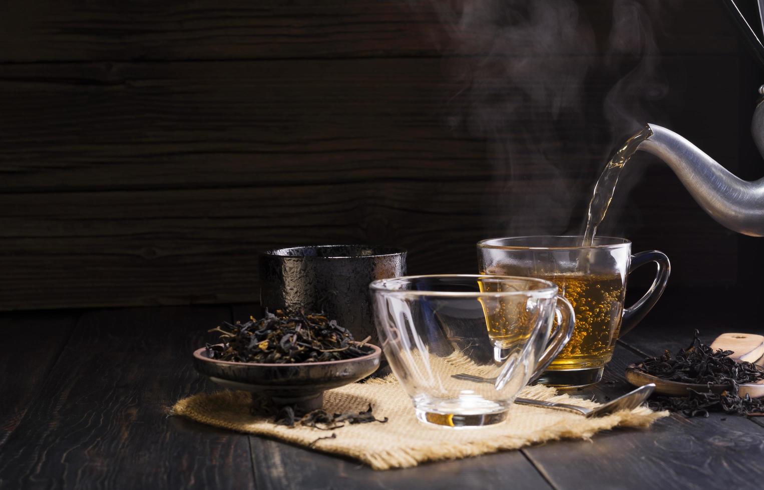 verser la théière chaude aux herbes dans une tasse en verre. la tasse de thé est placée sur une table en bois noire sur fond sombre. la lumière du soleil du nouveau matin brille dans l'atmosphère chaleureuse. photo