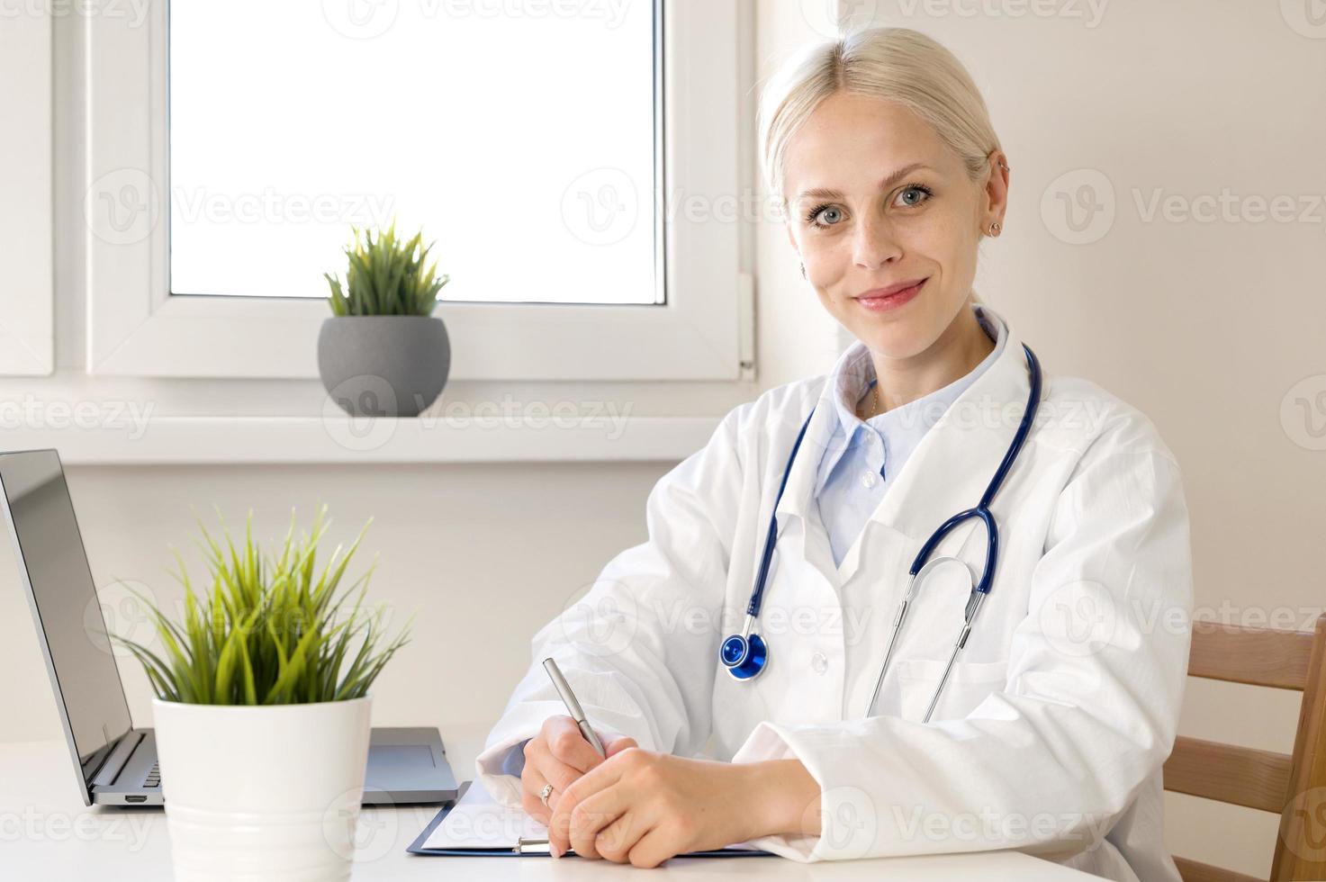 portrait en gros plan d'une jeune femme médecin heureuse souriant à la caméra assise dans son cabinet en notant l'historique des patients. photo