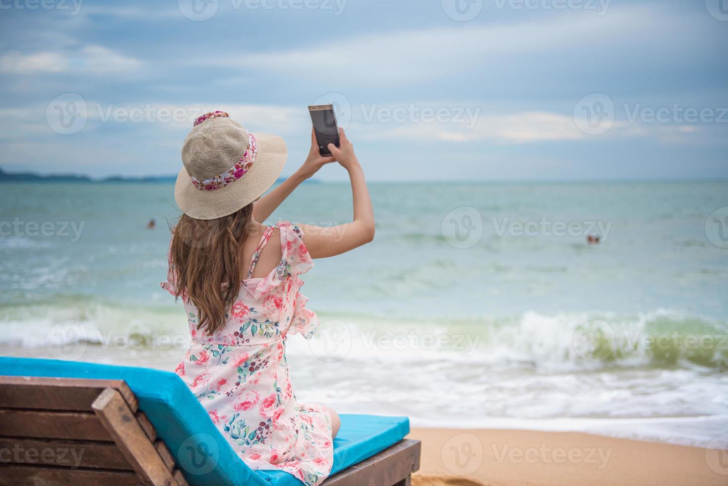 concept de voyage de vacances de vacances à la plage d'été, jeune femme asiatique heureuse avec un chapeau se relaxant et prenant une photo et un selfie avec un smartphone sur une chaise de plage.