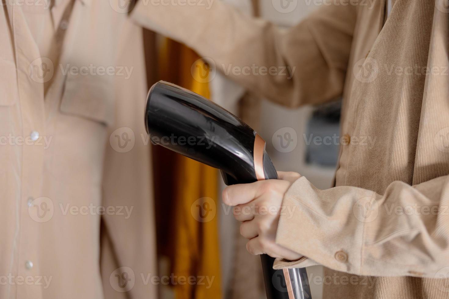 jeune femme fumant ses vêtements à la maison, vue rapprochée. femme utilisant un cuiseur à vapeur électrique, repassant des vêtements sur un cintre. technologies modernes. photo