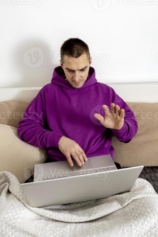 jeune homme caucasien avec sweat à capuche violet assis sur le lit et tenant un ordinateur portable. homme utilisant un ordinateur portable pour étudier en ligne, appeler des amis. enseignement à distance, appel vidéo, dire bonjour. détente à la maison. photo