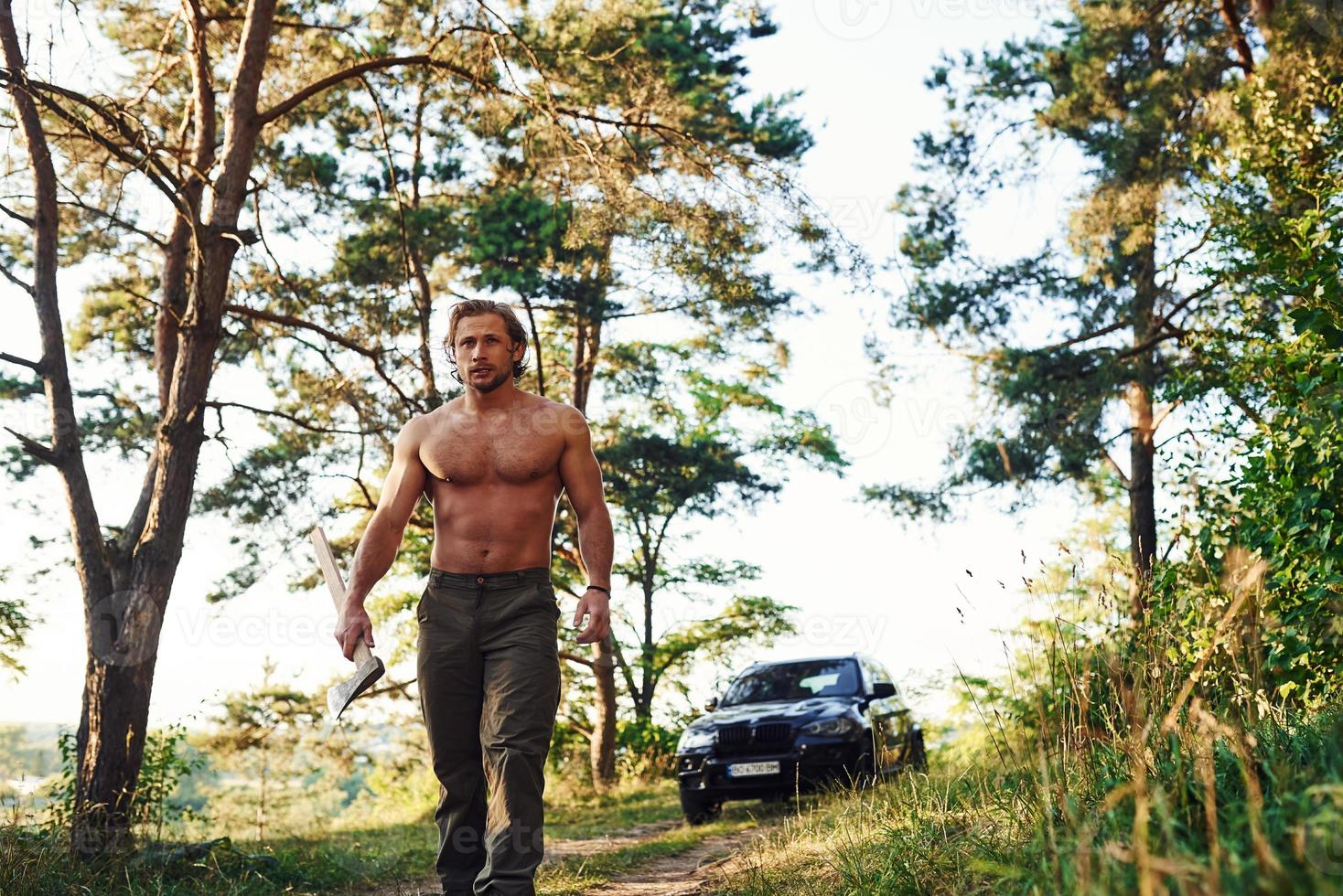 avec hache. bel homme torse nu avec un type de corps musclé est dans la forêt pendant la journée photo