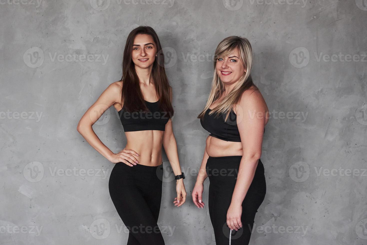 deux amies avec différents types de corps et en vêtements de sport debout dans le studio avec un fond gris photo