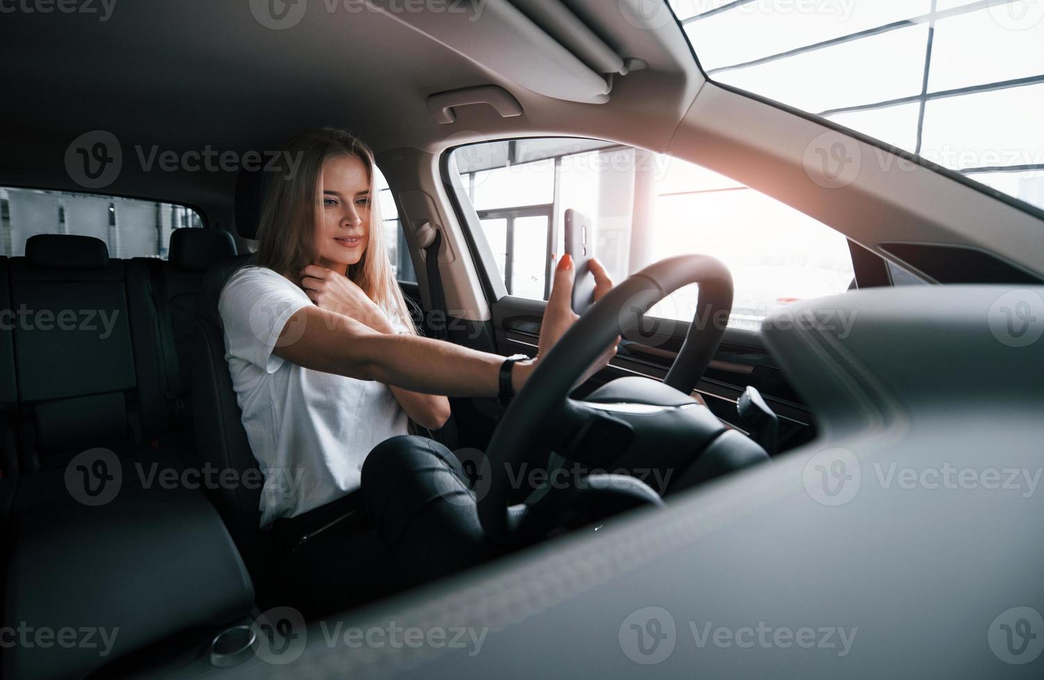 se regarde en utilisant la caméra frontale du smartphone. fille en voiture moderne dans le salon. la journée à l'intérieur. achat d'un véhicule neuf photo