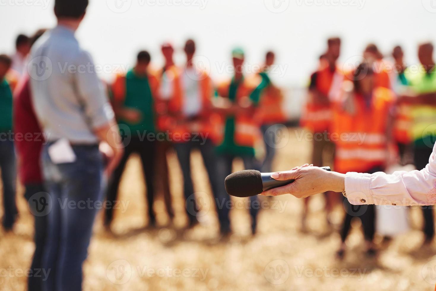 la main de la femme tient le microphone. un groupe de personnes en uniforme jaune se tient à l'extérieur sur le terrain photo