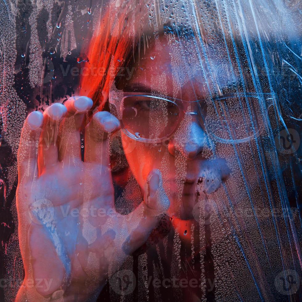 vue de face. studio tourné en studio sombre avec néon. portrait d'homme sérieux derrière la vitre humide photo