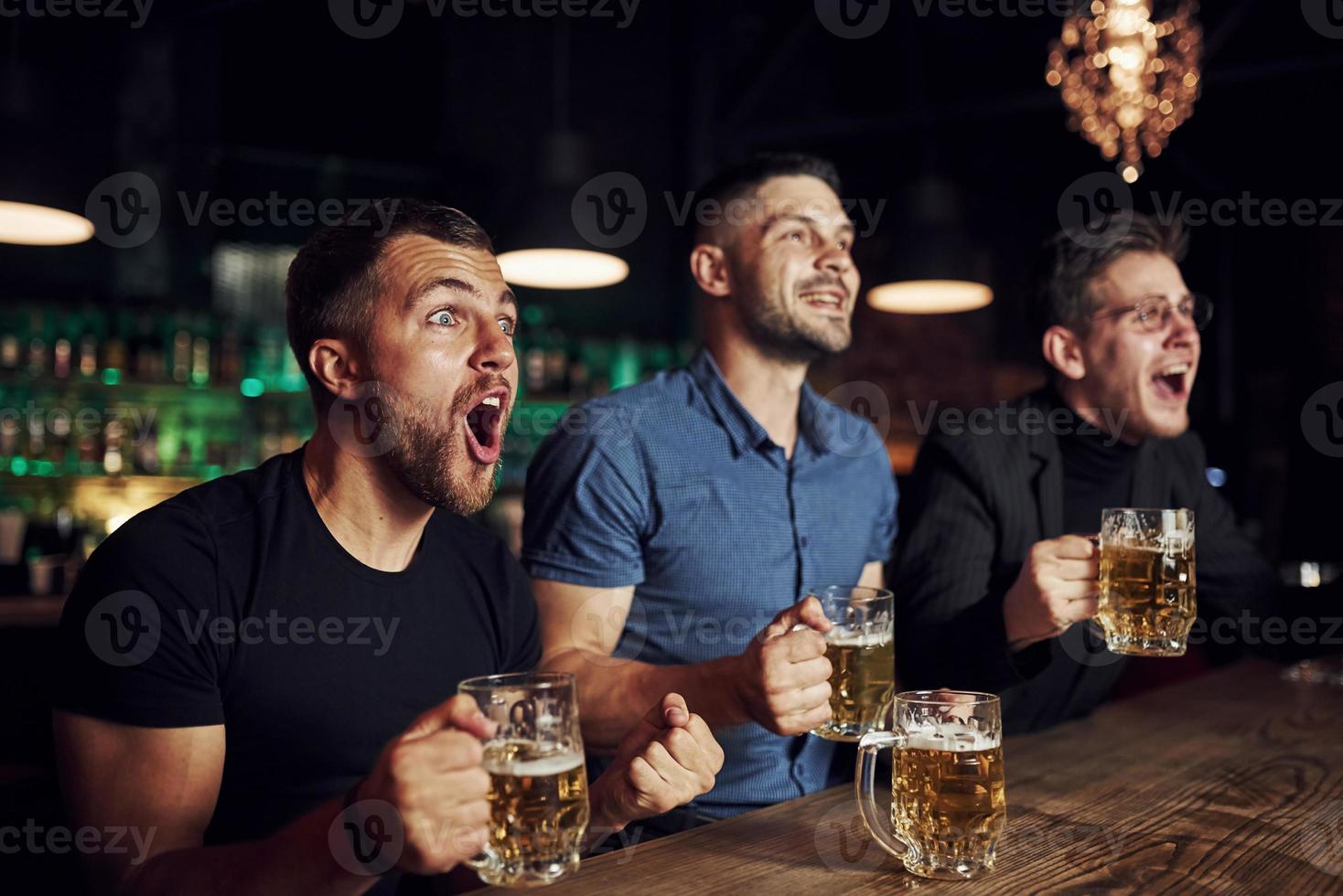 célébrant la victoire. trois fans de sport dans un bar regardent le football. avec de la bière dans les mains photo