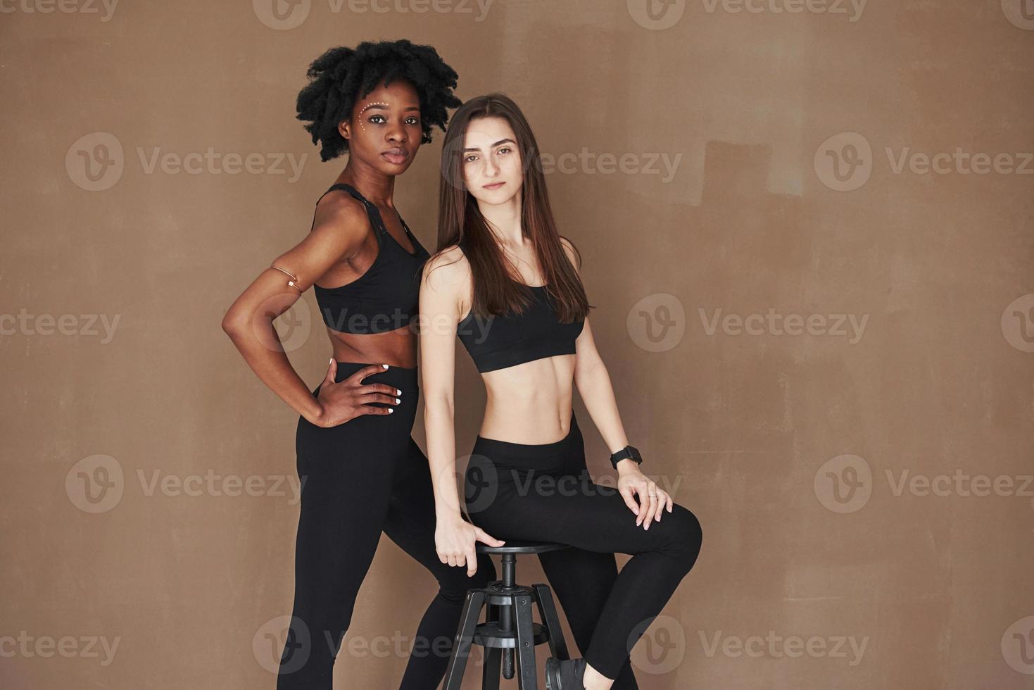 sur la chaise noire. deux amies multiethniques se tiennent dans le studio avec un fond marron photo