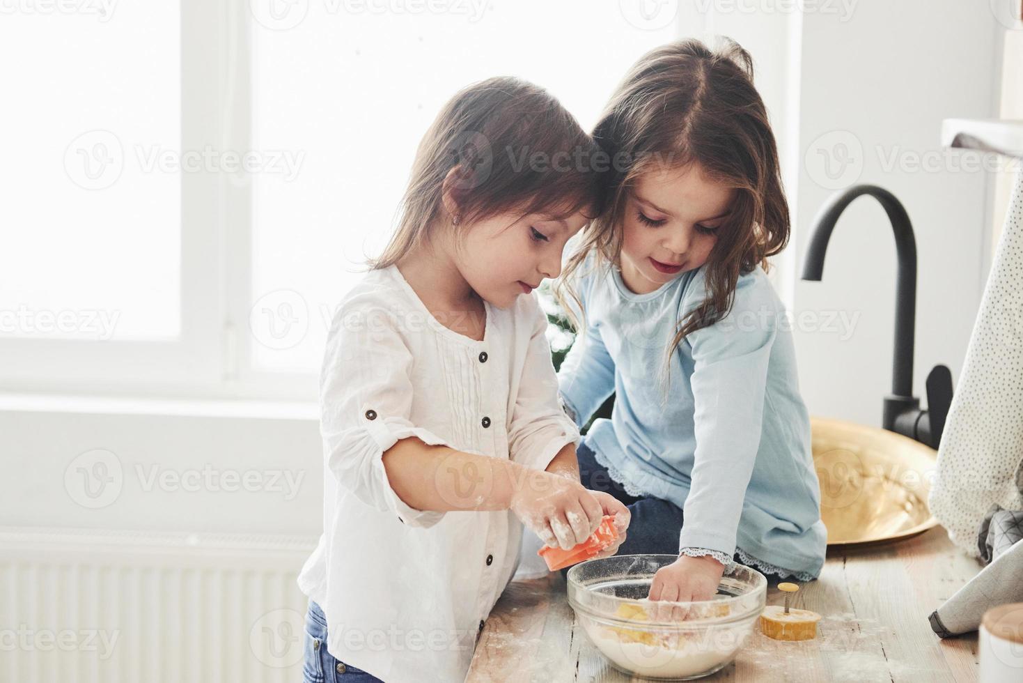 plus comme ça qu'ils jouent. amis d'âge préscolaire apprenant à cuisiner avec de la farine dans la cuisine blanche photo