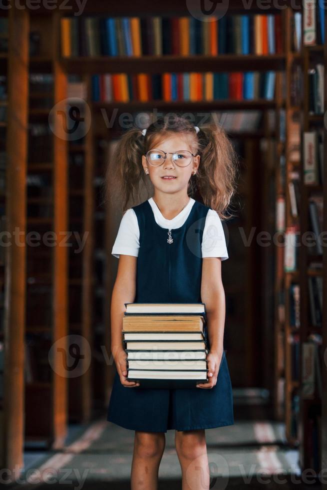 jolie petite fille à lunettes se tient dans la bibliothèque pleine de livres. conception de l'éducation photo