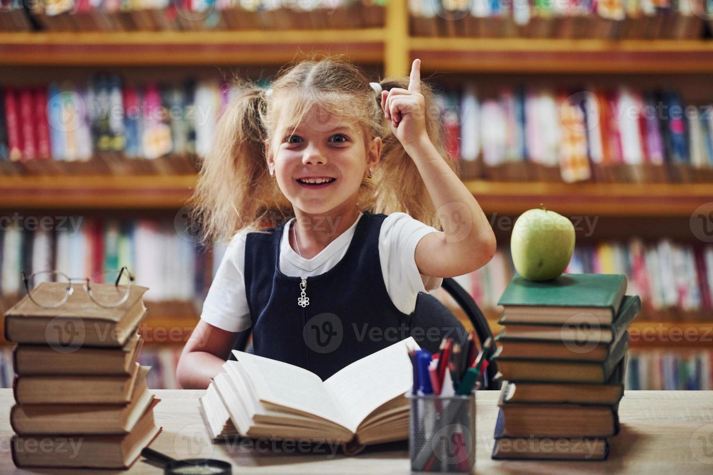 j'ai une idée. jolie petite fille avec des nattes est dans la bibliothèque. pomme sur les livres photo
