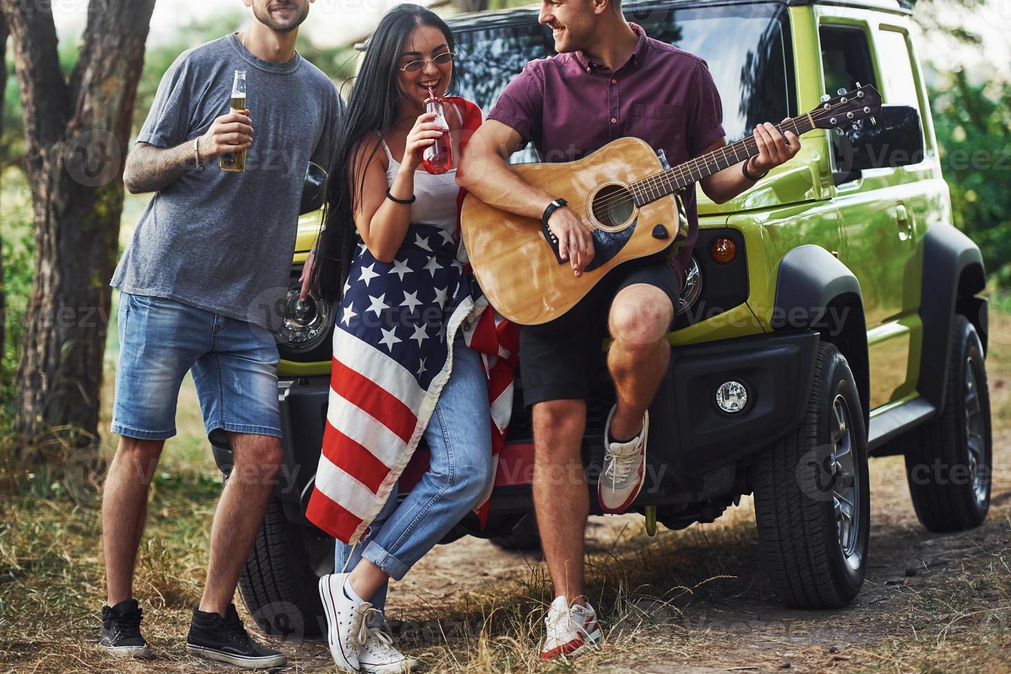 musicien joue une chanson à la guitare. les amis passent un bon week-end à l'extérieur près de leur voiture verte avec le drapeau américain photo