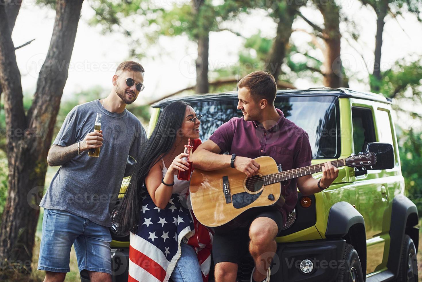 musicien joue une chanson à la guitare. les amis passent un bon week-end à l'extérieur près de leur voiture verte avec le drapeau américain photo