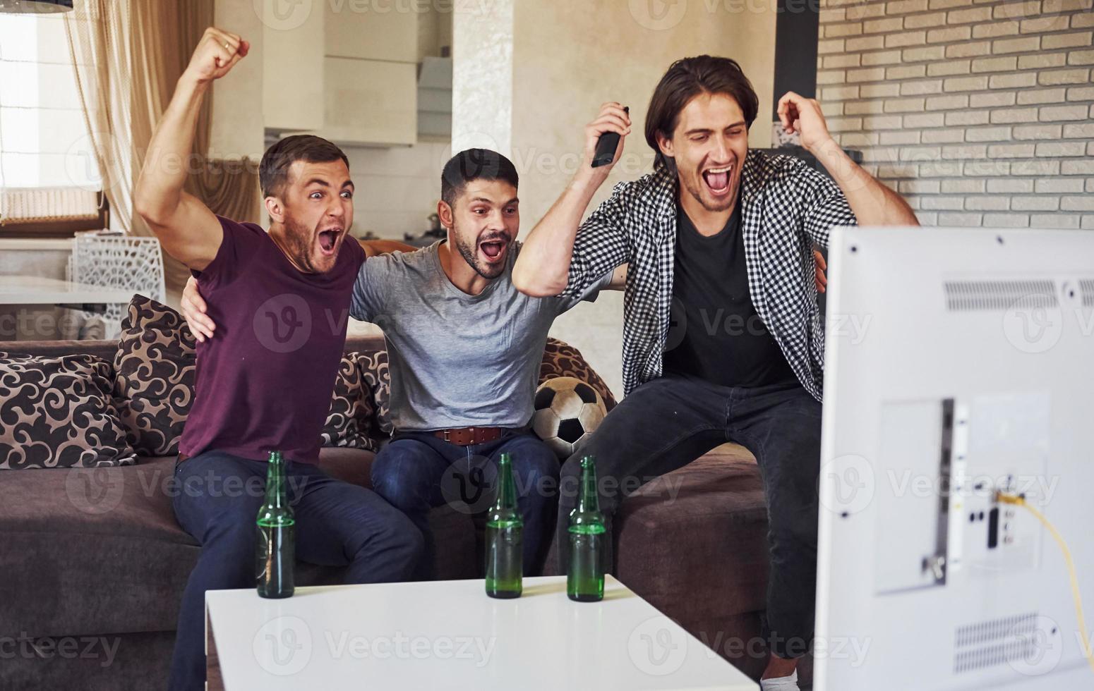 c'est un but. excité trois amis regardant le football à la télévision à la maison ensemble photo