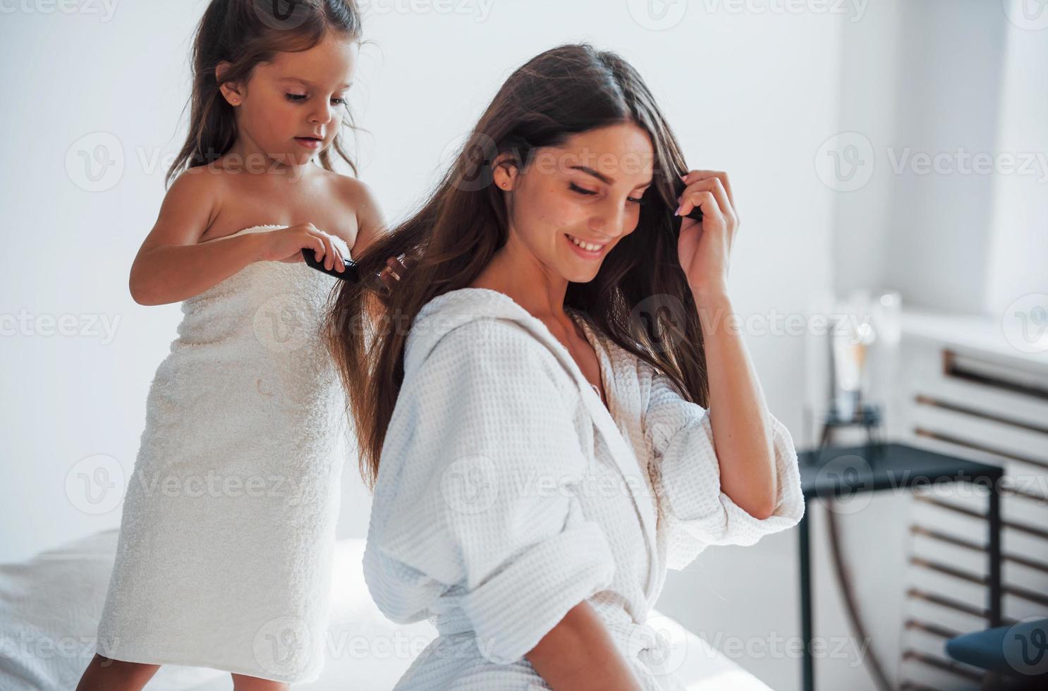 aider à faire la coiffure. jeune mère avec sa fille a une journée de beauté à l'intérieur dans une salle blanche photo