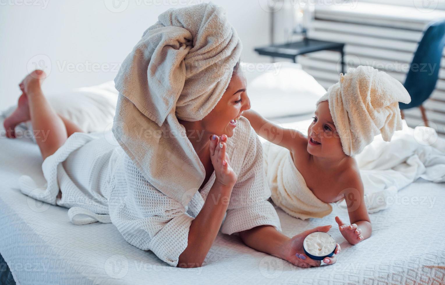 à l'aide de crème. jeune mère avec sa fille a une journée de beauté à l'intérieur dans une salle blanche photo