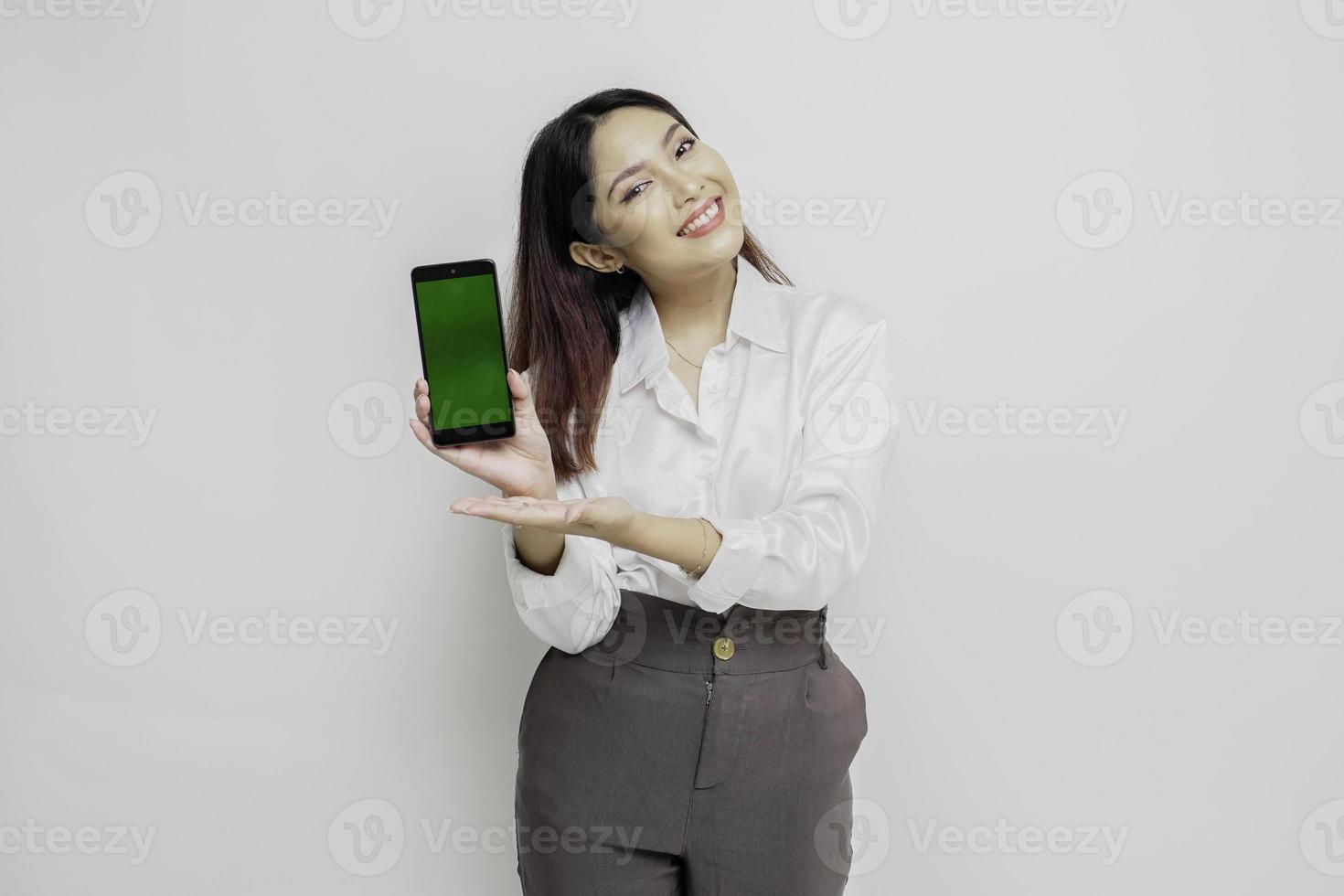 femme asiatique excitée portant une chemise blanche pointant vers l'espace de copie à côté d'elle tout en tenant son téléphone, isolée par fond blanc photo