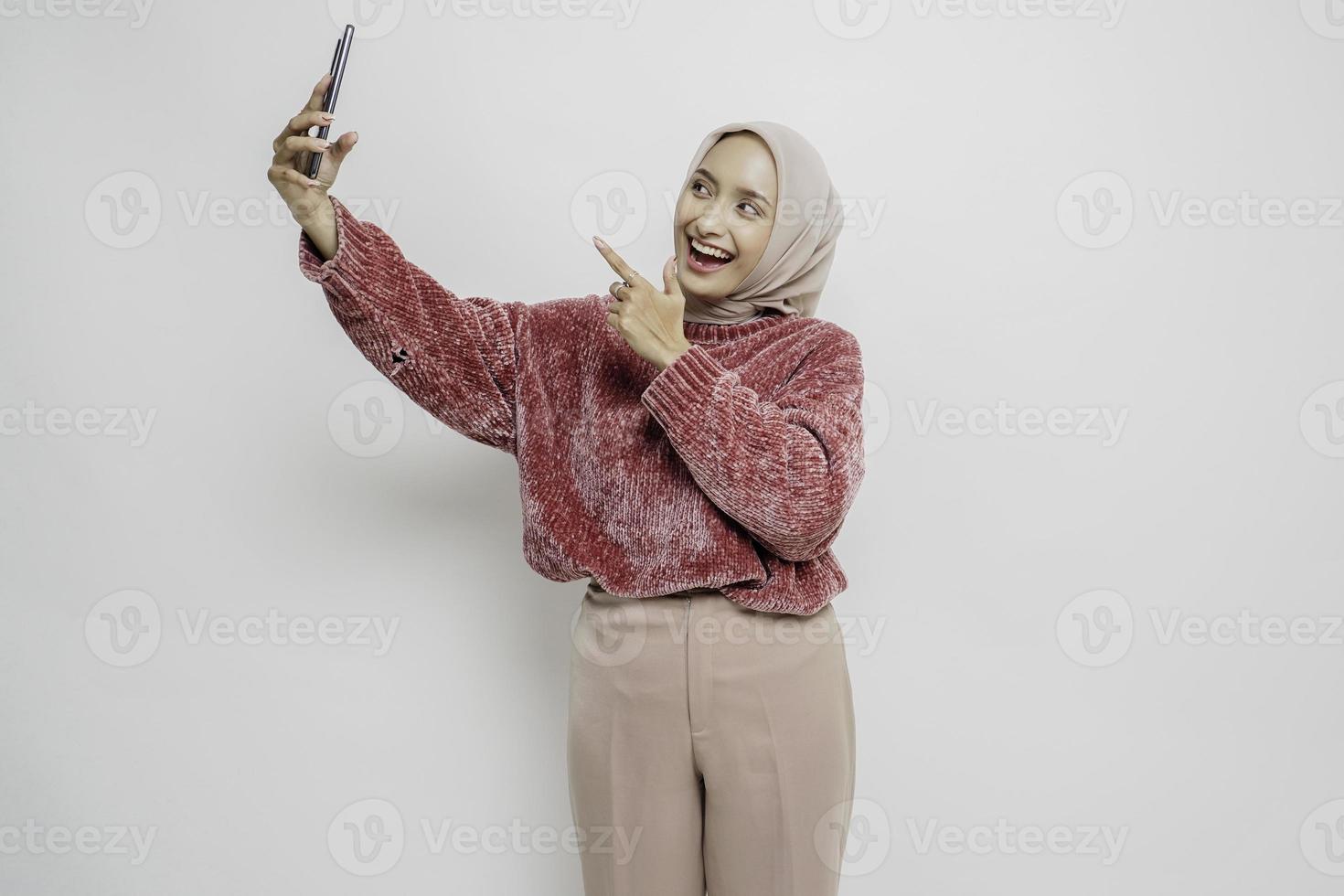 portrait d'une femme asiatique heureuse portant un chandail rose et un hijab, tenant son téléphone, isolée sur fond blanc photo