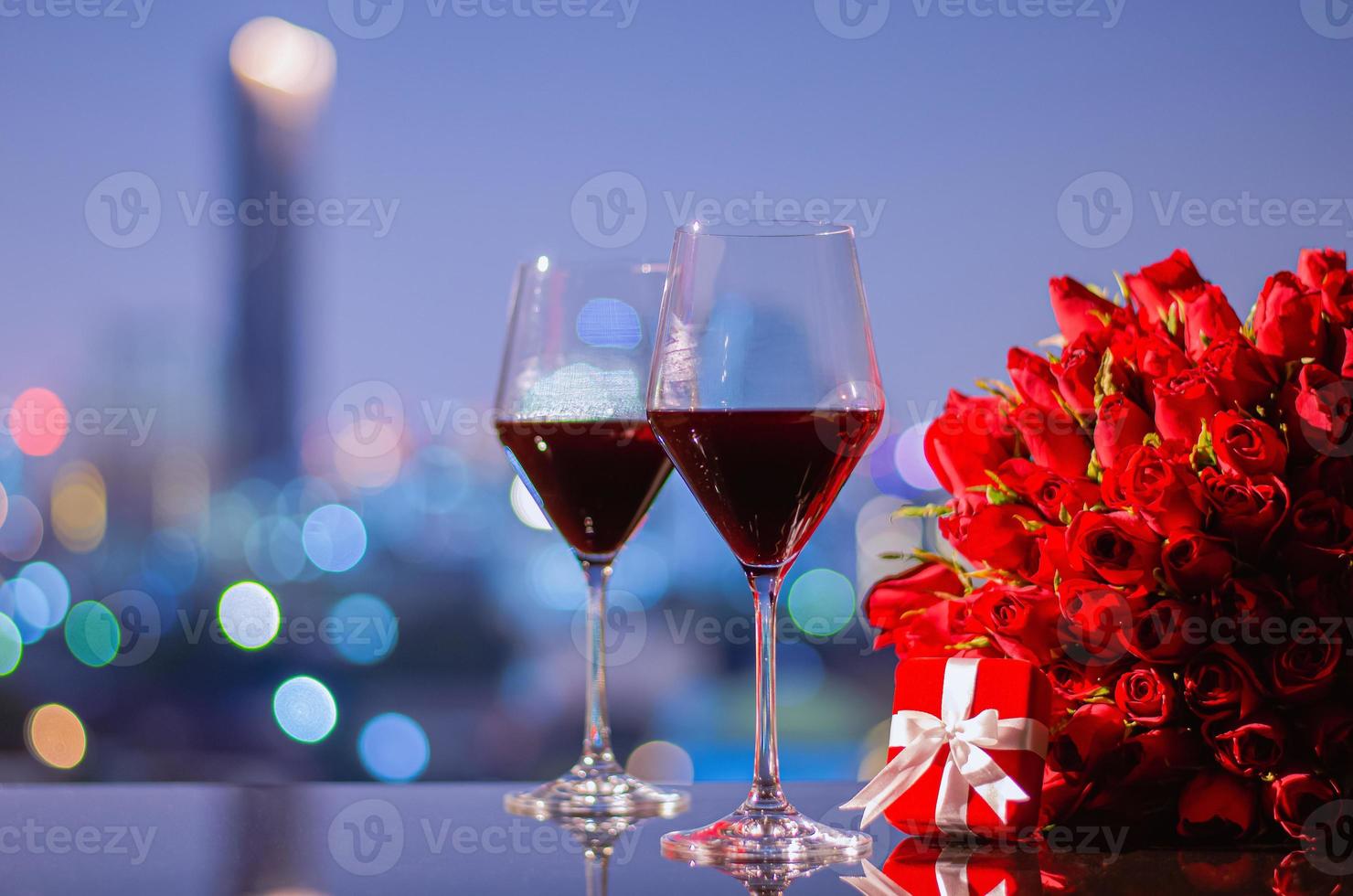 deux verres de vin rouge et bouquet de roses rouges avec boîte-cadeau rouge sur table avec lumières bokeh colorées de la ville pour le concept d'anniversaire ou de la saint-valentin. photo