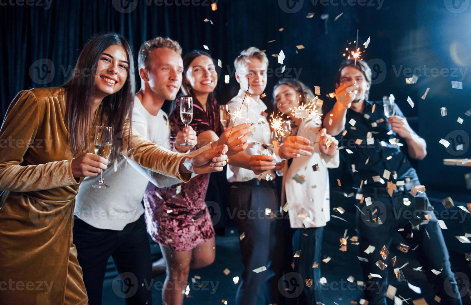 s'amuser avec des cierges magiques. les confettis sont dans l'air. groupe d'amis joyeux célébrant le nouvel an à l'intérieur avec des boissons dans les mains photo