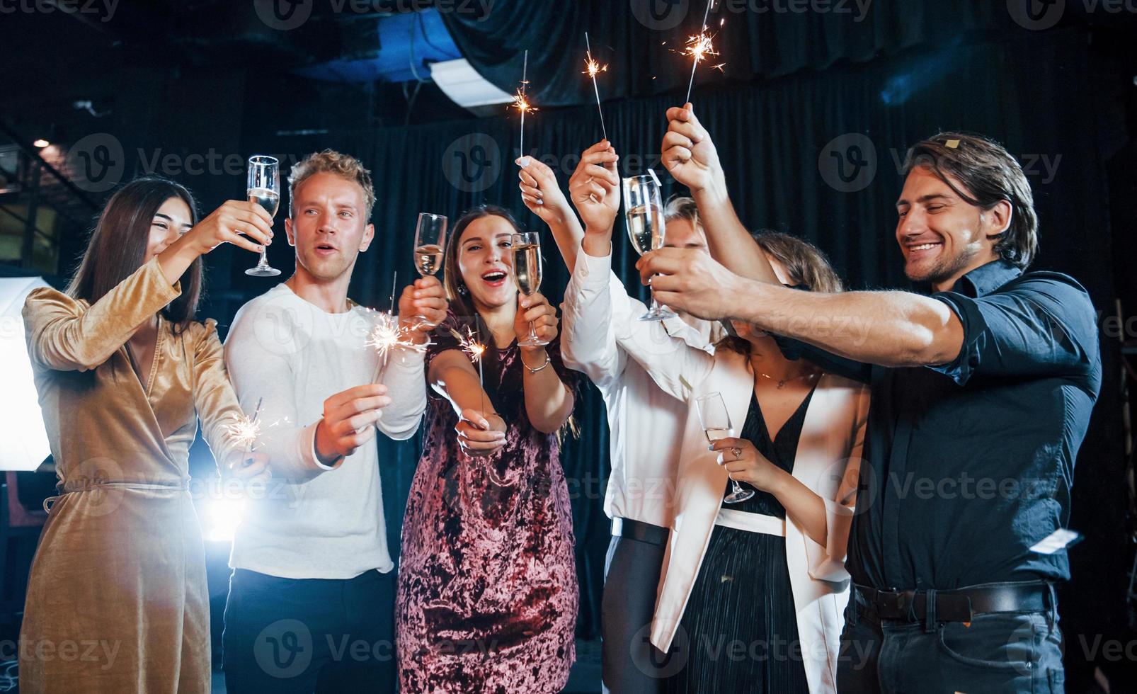 s'amuser avec des cierges magiques. groupe d'amis joyeux célébrant le nouvel an à l'intérieur avec des boissons dans les mains photo