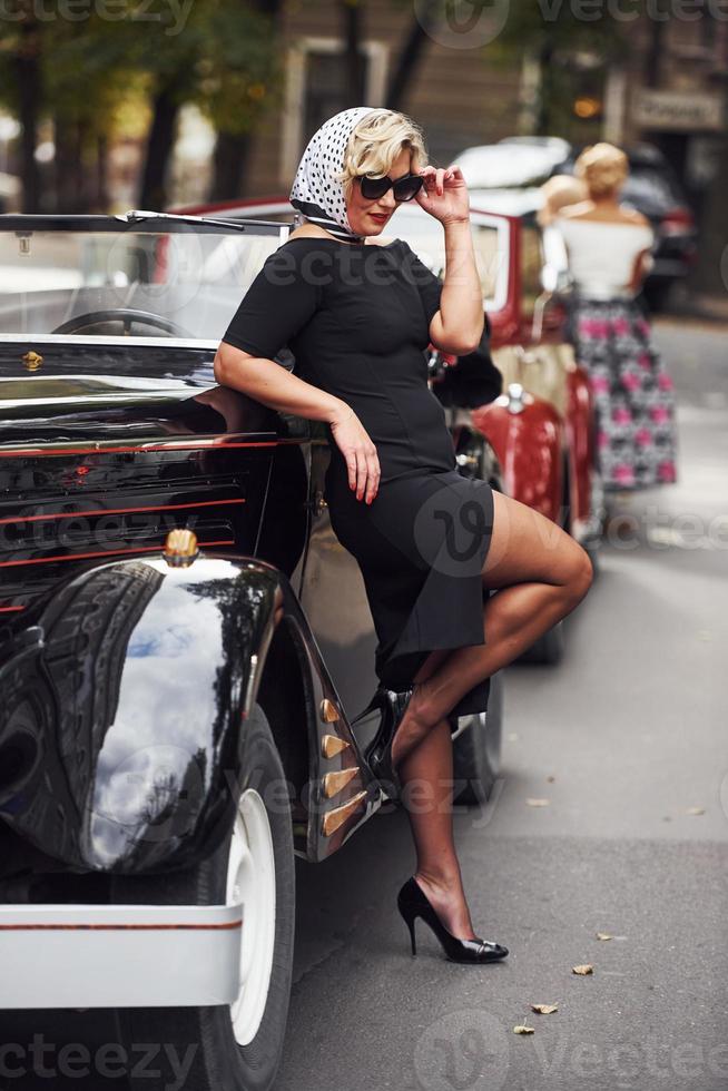 femme blonde à lunettes de soleil et en robe noire s'appuyant sur une vieille voiture classique vintage photo