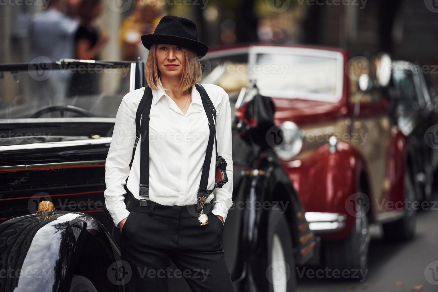 femme blonde au chapeau et en vêtements rétro noirs près de la vieille voiture classique vintage photo