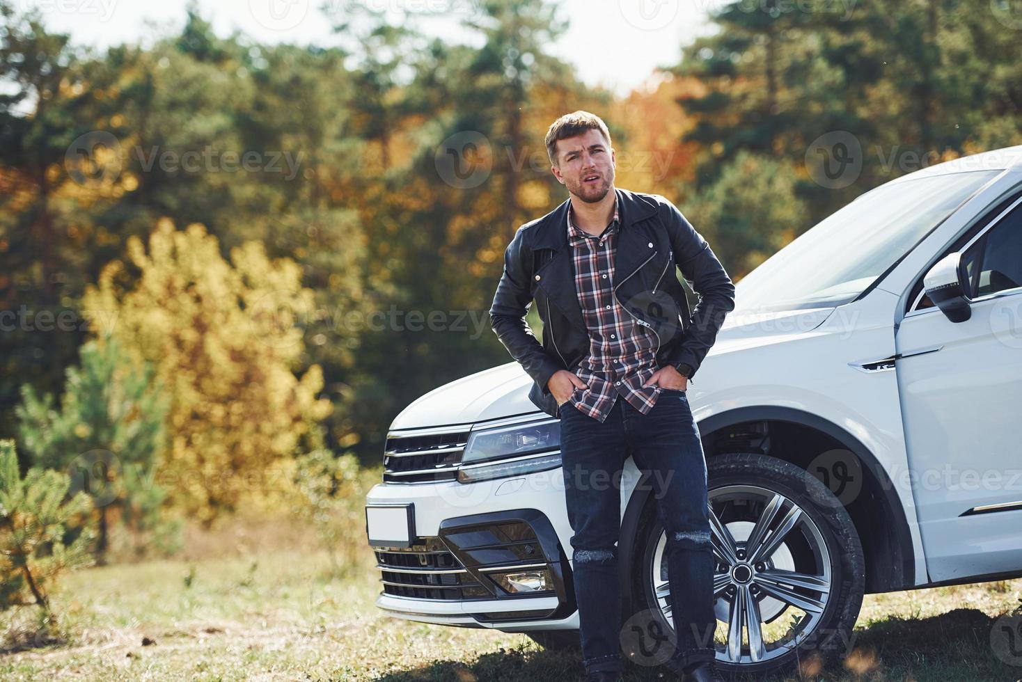 un homme en veste de cuir noir se tient près de sa voiture blanche garée à l'extérieur et attend de l'aide photo