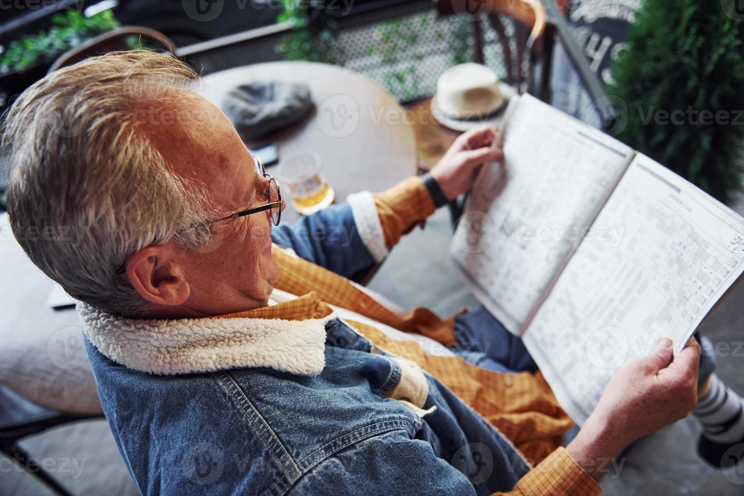 senior élégant dans des vêtements à la mode et des lunettes est assis dans le café et lit le journal photo