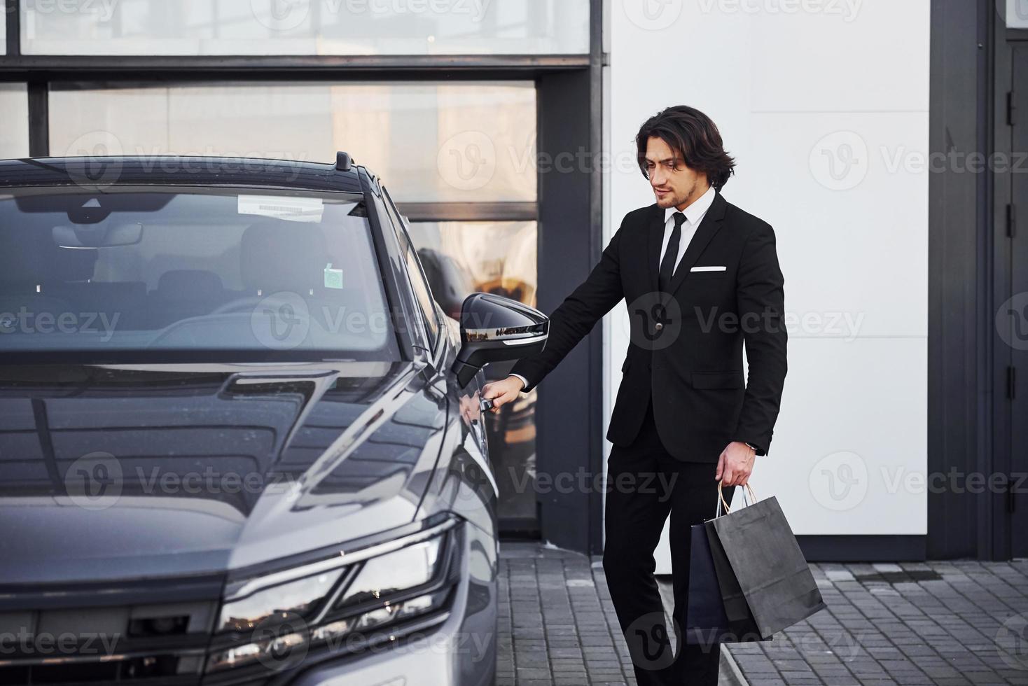 portrait d'un beau jeune homme d'affaires en costume noir et cravate à l'extérieur près d'une voiture moderne et avec des sacs à provisions photo