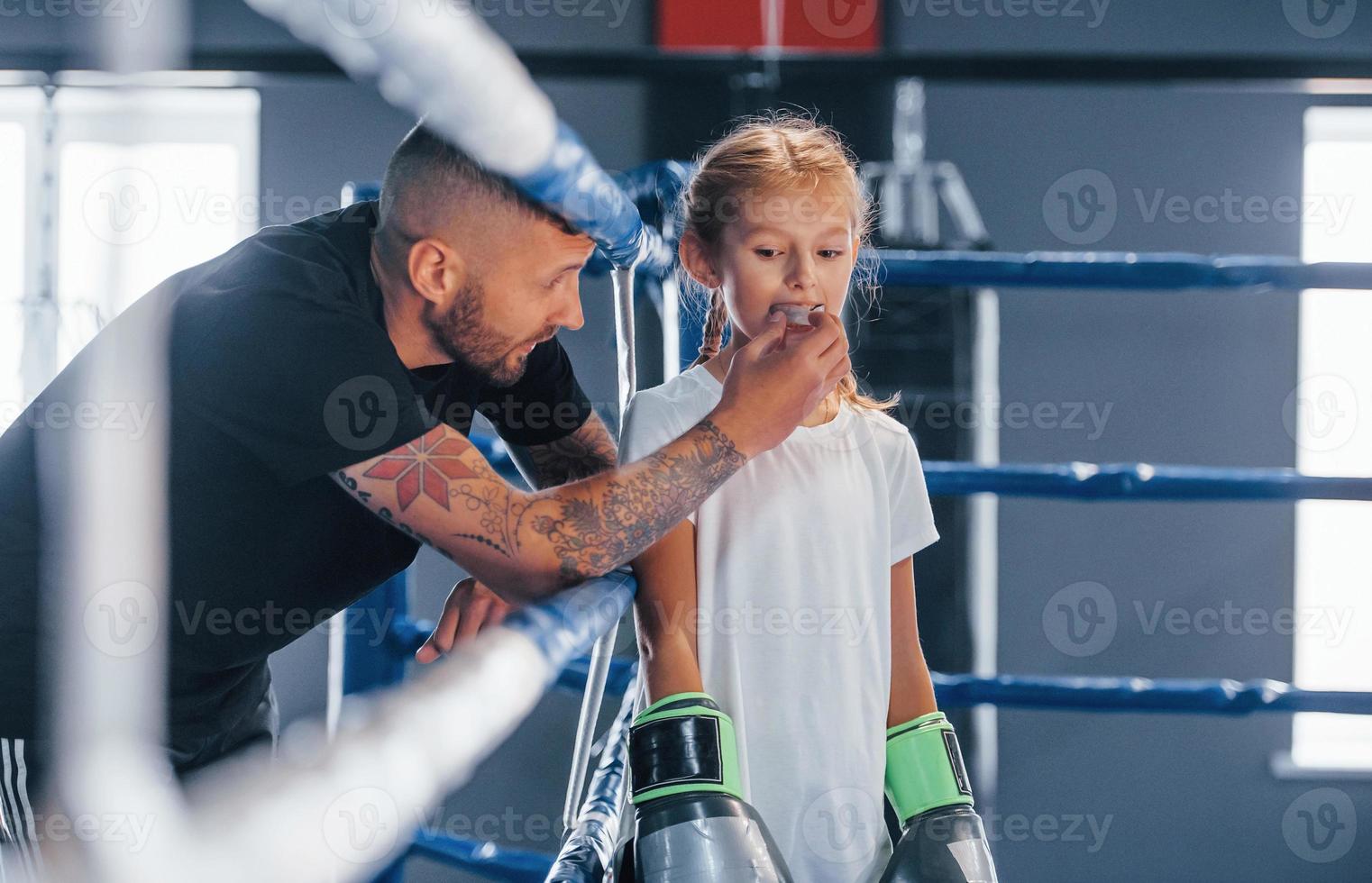 debout sur le ring de boxe. jeune entraîneur de boxe tatoué enseigne une jolie petite fille dans la salle de gym photo