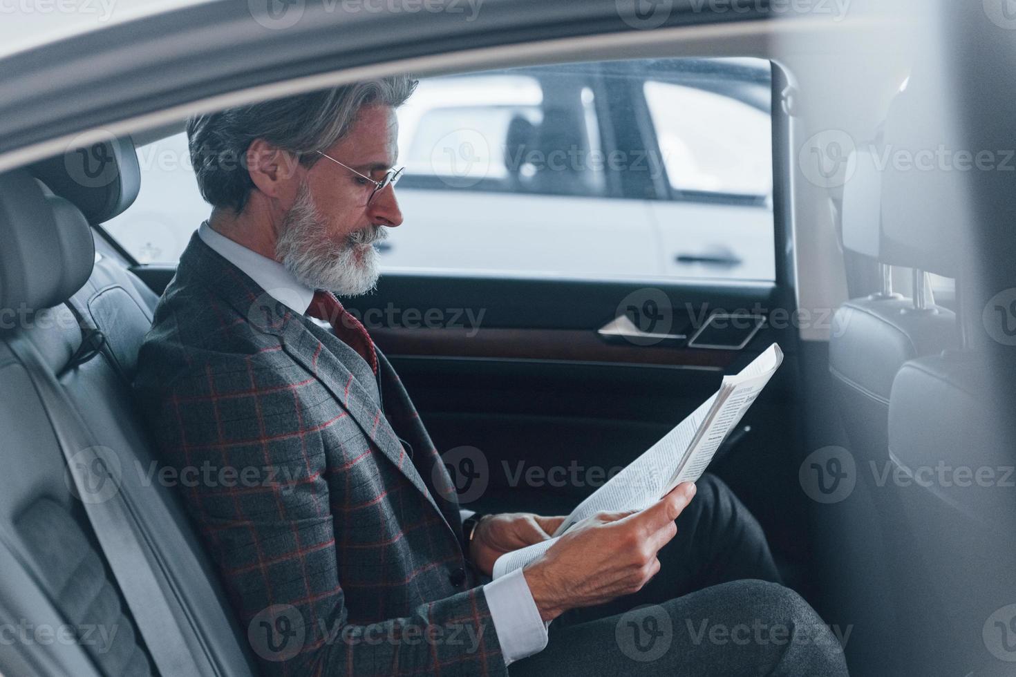 homme âgé moderne et élégant aux cheveux gris et à la moustache lisant le journal à l'intérieur de la voiture photo