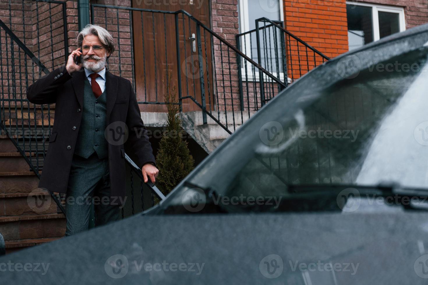 un homme âgé à la mode aux cheveux gris et à la barbe a une conversation par téléphone à l'extérieur dans la rue près de sa voiture photo