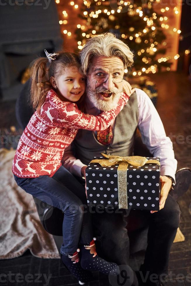 homme âgé à la mode joyeuse avec des cheveux gris et une barbe assis avec une petite fille dans la salle de noël décorée photo