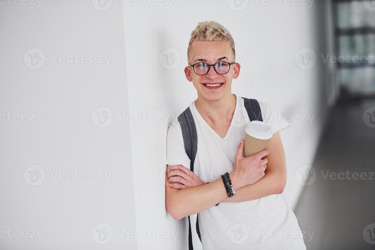 étudiant en vêtements décontractés et avec sac à dos se tient à l'intérieur contre un mur blanc avec une tasse de boisson photo