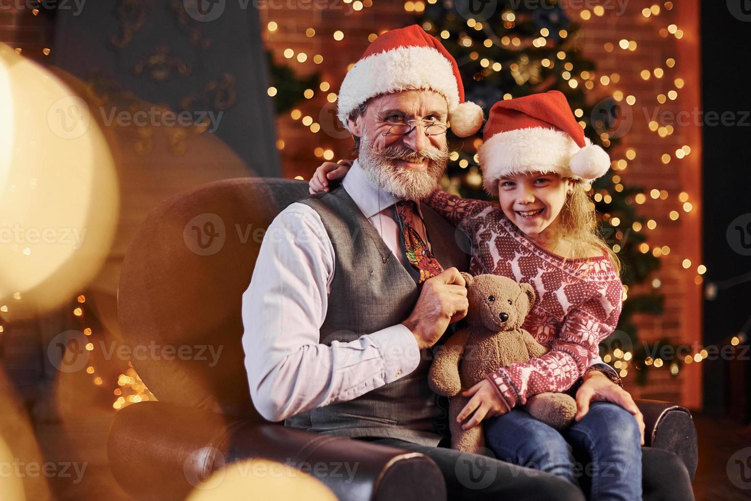 homme senior à la mode joyeuse avec des cheveux gris et une barbe assis avec une petite fille dans des chapeaux de noël et un ours en peluche photo